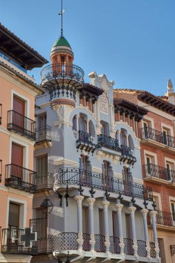 Teruel 'deki El Torico Tekstil Evi, tarihî binalarla çevrili yumuşak tonlarıyla ve dövülmüş demir balkonlarıyla modernist cepheleriyle göze çarpıyor. Günbatımının sıcacık ışığı süssel detayları ve dekoratif kubbeyi vurgular.