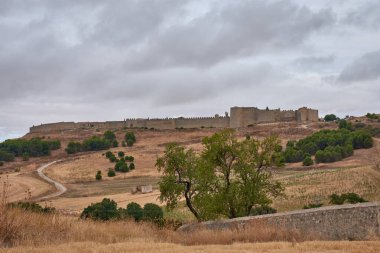 Panoramic view of the walled city of Uruena, in Valladolid, Castile and Leon, Spain, with its imposing medieval stone wall stretching along a hill. The surrounding landscape of dry fields and sparse vegetation under a dramatic