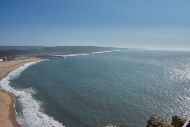 Yüksek bir bakış açısından, yeşil tepeler ve beyaz mimari ile çevrili kıyıları koruyan eğimli limanın yanındaki Nazare altın plajını görebilirsiniz. Mavi deniz ve berrak gökyüzü Sahneye derinlik ve dinginlik katar. 