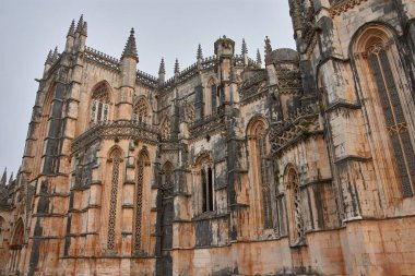 Batalha Manastırı, karmaşık Gotik süslemeleri ve Kılavuz unsurları ile sivri kemerleri, iz sürme yerleri ve siluetini tanımlayan zirveleriyle. Yaşlanmış taş ve iskeleli alanlar, tarihsel değer ve koruma süreçlerini vurguluyor.