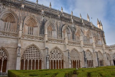 From the inner courtyard of the cloister of the Batalha Monastery, the Gothic facade of the cathedral can be seen, with elaborate tracery, pinnacles, and pointed arches rising above a symmetrical garden. 
