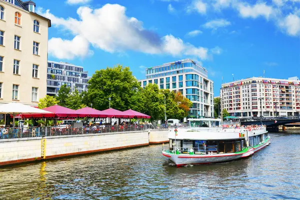 Berlin, Almanya - 31 Temmuz 2024 - Fotoğraf Spree 'de bir turistik gezi teknesi, bir köprü ve arka planda çeşitli yerleşim binalarını gösteriyor.