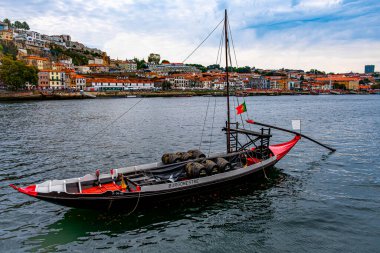 Porto şehrinin arka planında, turuncu çatılı ve kasvetli havalarda evleri olan kırmızı tekne nehirde yüzüyor.