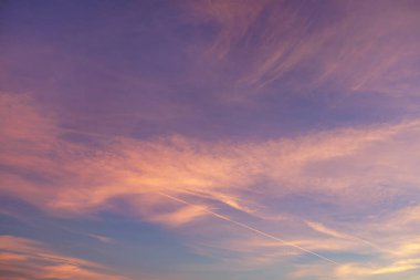 A serene sky transitions from soft blue near the horizon to shades of pink and purple as it reaches higher into the sky. Wispy clouds and a faint diagonal contrail create a dynamic yet tranquil scene.