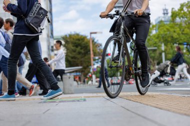 Bir bisikletçi ve birkaç yaya parçalı bulutlu bir gökyüzünün altında kalabalık bir şehir caddesinden geçiyorlar. Düşük açı ve yakın çekim fotoğrafı resmin hareketli atmosferine eklenir.