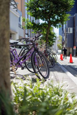 Bisikletler Nagoya 'da ağaçlar ve modern tasarımın binalarıyla kaplı güneşli bir şehir caddesi boyunca park halindedir. Parlak turuncu trafik konileri olan bir perosn yan sokakta yürüyor..