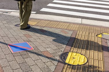 Uzun gölgeler ve bol kargo pantolonları, Japonya 'da zebra soyulmuş yaya geçidine daha iyi erişebilmek için tasarlanmış dokunsal asfalt döşeme üzerinde duran genç bir kadının yer aldığı şehir sahnesi için odak noktası oluşturuyor..