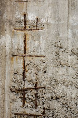Paslı inşaat demiri ızgarası bitmemiş, yıpranmış beton bir duvardan dışarı çıkar ve duvarın monoton gri dokusuna turuncu kahverengi renkler ekler ekler. Beton karışımına eklenen küçük taşlar da görülebilir..