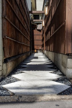 Yüksek ahşap duvarlarla çevrili dar yol kırsal Japon restoranının görünmeyen girişine çıkıyor. Kömür grisi kayalarla çevrili, güneş ışığı ve gölgelerle aydınlatılmış desenli taş yollar..