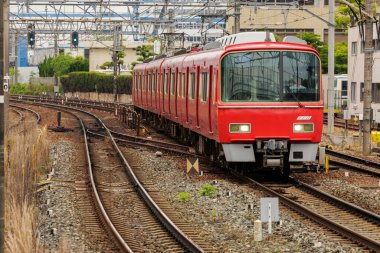 Parlak kırmızı tren, binalar, elektrik hatları ve sinyallerle çevrili kavisli bir hat boyunca ilerliyor. Demiryolu çevresinin sessiz tonlarıyla trenin parlak tasarımı ve canlı renk kontrastı.