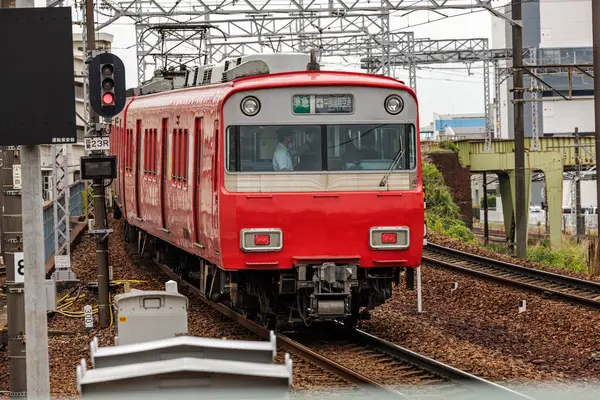 Kıvrımlı raylarda parlayan parlak kırmızı tren peronundan çıkarken yakın plan, arka vagonun penceresinden antreman şefi görünür. Tepedeki elektrik hatları ve demiryolu altyapısı çerçevesi.