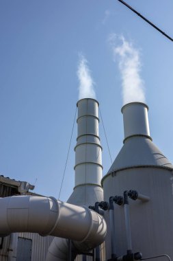 Uzun sanayi bacaları açık mavi gökyüzüne beyaz buhar yayar. Sanayi altyapısı ve çevresel etki temalarını gösteriyor. Önplanda büyük borular ve silindirik yapılar öne çıkıyor.