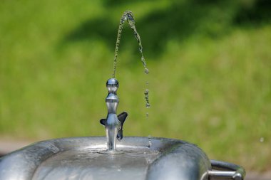 Krom renkli musluktan fışkıran su yayı ile yakın plan fıskiyeler. Güneş ışığı, çağlayan su damlacıkları Bokeh yeşilliğine düşerken akıntının berraklığını vurgular.