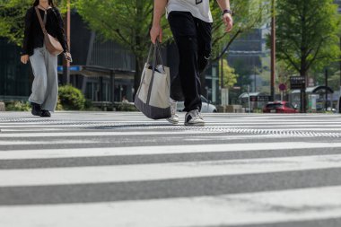 Alçak açılı kentsel yaya geçidi. Gündelik giysiler giymiş iki genç beyaz zebra çizgileriyle kesişiyor. Odaklanma ön planda, spor ayakkabılı, çantalı bir erkek..