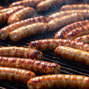 Izgarada cızırdayan çoklu sosislerin göz hizasında makro fotoğraf, her bir sosis meyve suyuyla parıldıyor, arka plandaki Oktoberfest kalabalığını bulanıklaştıran sığ odak