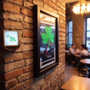 shamrock picture on craft paper in frame hanging on brick wall in the pub, blurred people drinking beer on background clipart