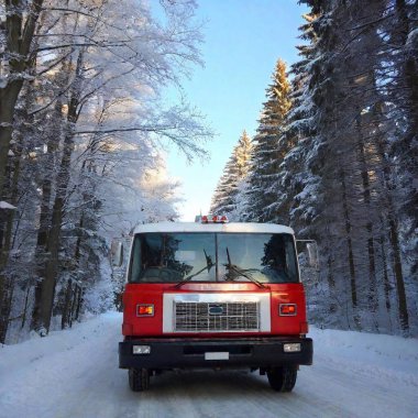 The photo shows a fire truck driving through a snowy winter landscape. The picture is taken from a low angle near the front wheel, which is partially covered in snow and ice. A chunky tire can be seen in the foreground, while a snow-covered road clipart