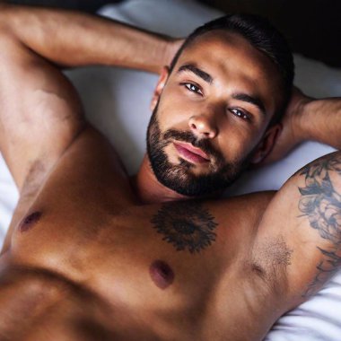 A high-resolution ultrarealistic image of a shirtless, muscular man with intricate tribal tattoos on his shoulder and upper arm, lying on a bed. The image is a photograph, capturing fine details of his tattoos, skin texture, and facial expression clipart