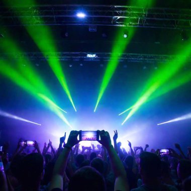 A high-resolution ultrarealistic photo capturing a vibrant concert scene. The image focuses on a large crowd with hands raised, illuminated by vivid green, purple, and blue laser lights cutting through the air. The stage features an energetic clipart