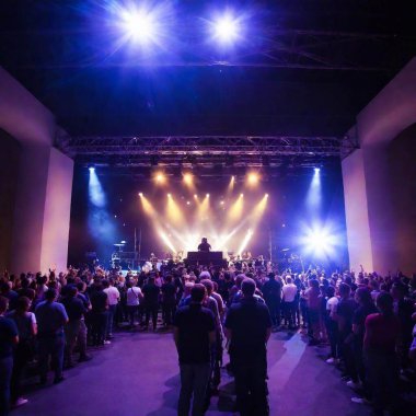 A high-resolution photo captures a large outdoor music concert at night. The stage, adorned with bright spotlights in hues of blue, white, and red, illuminates the performing band. The audience, seen as silhouettes in the foreground, faces the stage clipart