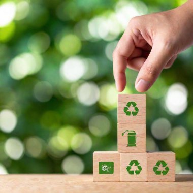 A high-resolution ultrarealistic image depicting a close-up of a hand stacking wooden blocks, each block displaying eco-friendly icons. The hand is in the process of placing the top block that features a recycling symbol. Other blocks include clipart