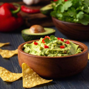 A high-resolution ultrarealistic image of delicious guacamole in a dark wooden bowl, garnished with fresh cilantro and diced tomatoes. The creamy, textured avocado dip is vibrant green, contrasting with the red pepper and lime in the background clipart