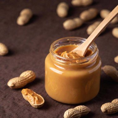 A high-resolution ultrarealistic image of a jar of creamy peanut butter placed on a brown woven fabric surface. The jar is open, revealing its smooth, shiny surface, and positioned to the right of the composition. Resting on top of the jar is a clipart