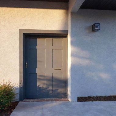 A high-resolution ultrarealistic image depicting the entrance of a modern residential home. This digital photograph features a sleek, charcoal-colored garage door occupying the left side, contrasted by smooth, light-grey walls. On the right, a clipart