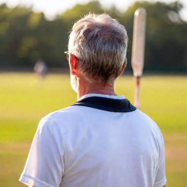 Caucasian senior male cricketer looking at the field, with the blurred stumps and fielders in the background. clipart