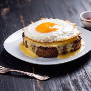 A crisp close-up of a croque madame with perfectly melted cheese and sunny-side egg on a clean white plate, the dark background adding a sophisticated touch. clipart