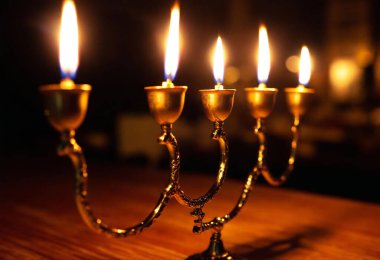 A side view of three golden menorah candles glowing brightly in front of traditional food, casting shadows on a wood table. clipart