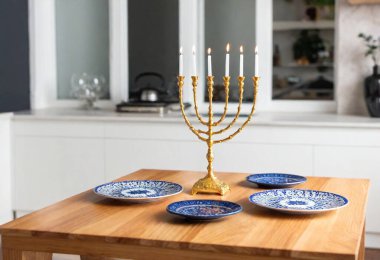 A full shot of a kitchen counter with a Hanukkah table setting featuring plates, gold flatware, and a brightly shining menorah. clipart