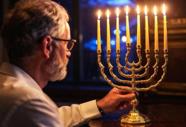 A back view of a male lighting the candles on the menorah, with reflections of the flames on a glass window beside him. clipart