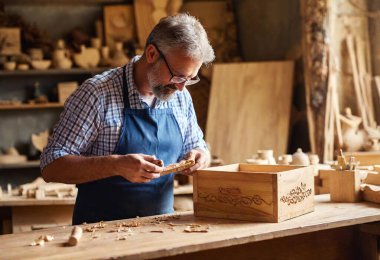 Woodworking Artist at Work: A cozy shop filled with the scent of sawdust, showcasing an artisan carving detailed patterns into a wooden box, with finished items on display nearby. clipart