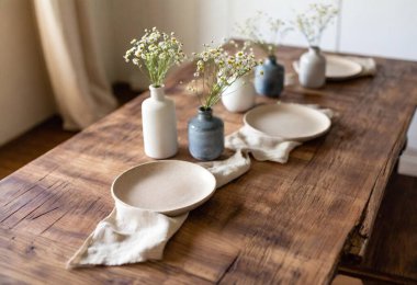 Casual setting with beige linen runners over a weathered wooden table, accented by handmade pottery plates, woven placemats, and small vases of daisies clipart