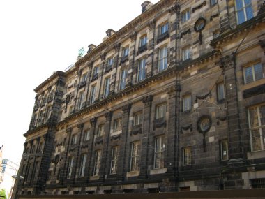 Görüntü, Amsterdam 'daki Baraj Meydanı' ndaki Kraliyet Sarayı 'nın kumtaşı gölgeleri, uzun pencereler ve merkezi bir kubbeyle büyük klasik mimarisinin açılı bir görüntüsünü sunuyor.