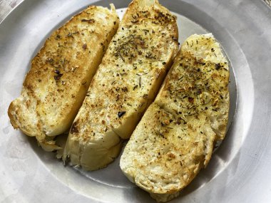 Üç altın-kahverengi sarımsaklı ekmek, pürüzsüz bir çelik tabağın üzerinde uzanır, kenarları çıtır çıtır ve tereyağlı..