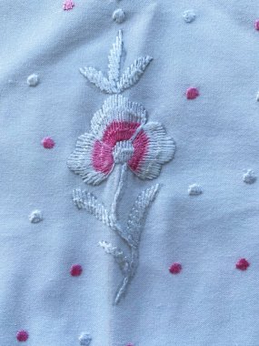 Yumuşak beyaz ve pembe ipliklerle işlenmiş ince işlemeli bir çiçek butta beyaz pamuk kumaşı süslüyor, ince işçilik sergiliyor.