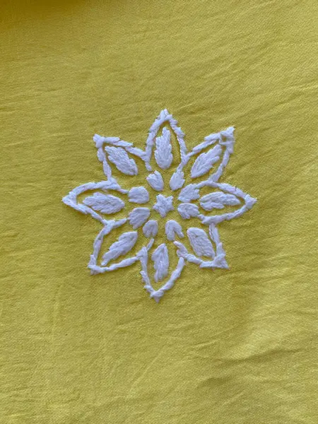 Zarif bir şekilde beyaz iplikle işlenmiş geometrik bir çiçek, parlak sarı ipek kumaşın üzerinde duruyor..