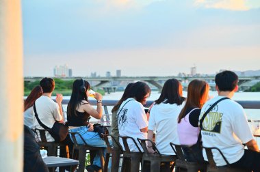 Tayvan - 28 Temmuz 2024: Turistler Dadaocheng İskelesi Plaza 'da büyüleyici bir günbatımının tadını çıkarırken, Taipei' de canlı bir nehir kenarı noktası olan satıcıların tezgahları ve gece aydınlatmaları ile turistlerin tadını çıkarıyorlar..