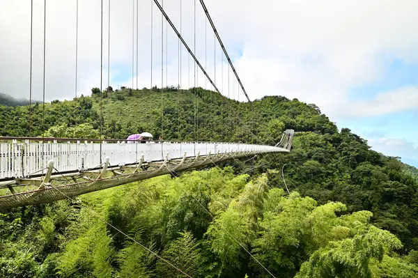 stock image Taiwan - Jul 07, 2024: Tourists stroll with umbrellas on Taiping Suspension Bridge under a clear summer sky, enjoying lush greenery and expansive natural views.