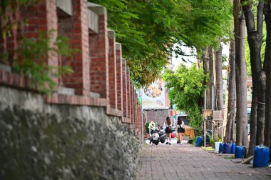 Taiwan - Jul 09, 2024: Two long-haired women in short skirts walk side by side along a red brick wall on a sunny day, with lush green trees adding a vibrant backdrop. clipart