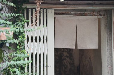 Solda yuvarlak yapraklı okaliptüslerle çevrili, solda ve sağda beyaz bir Japon noreniyle süslenmiş, sadelik ve zarafetin bir karışımını çağrıştıran huzurlu bir bahçe girişi..