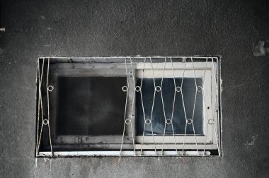 Yaşlanan bir beton duvar siyah toz taşır, beyaz boyalı ahşap bir pencereyi demir ızgaralarla çerçeveler. Yıllar süren yıpranma ve ihmalkârlığın kısmen kırılan ipuçları..