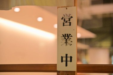 Beyaz duvarlar, sıcak ışıklandırma ve doğal güneş ışığının yer aldığı rahat bir yemek ortamı, cam kapıya odaklanmış bir asılma işareti 