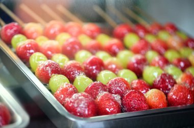 Çilek ve üzümlü bir tabak şekerli meyve şişi sergileniyor. Meyveler parlak bir şeker katmanıyla kaplıdır, arka plan şişleri vurgulamak için hafifçe bulanıklaşır..