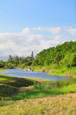 Tayvan 'ın Yilan kentindeki Zhuangwei Dune Ekoloji Parkı' nın sakin manzarası, kavisli mavi su gövdesi, yemyeşil çimenler, orman yamaçları, dağınık kayalar ve parlak mavi bir gökyüzü..