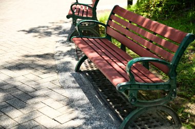 Kırmızı-kahverengi tahta levhalar ve süslemeli koyu yeşil çerçeveler içeren park bankları açık renkli tuğla patika boyunca konum çimen alanları ve tepedeki ağaçların gölge desenleri.