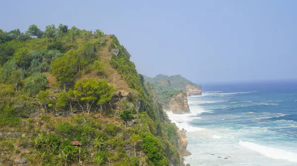 Sahildeki yüksek kayalıkların manzarası, mavi denizin güzelliği ve arka planda çarpıcı dalgalar. Seruni plajı, Yogyakarta, Endonezya