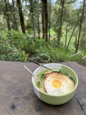 Ahşap masada yumurtalı ramen eriştesi ve arka planda doğa ormanı manzarası.